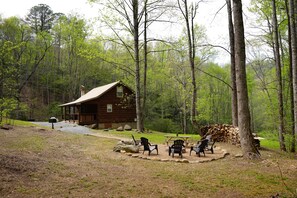A rustic cabin with a wooden exterior, surrounded by a forest. There are several Adirondack chairs arranged around a picnic table.