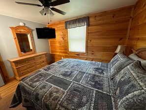A bedroom with a wooden wall, a bed with a quilt, a dresser, a TV, and a window with blinds.