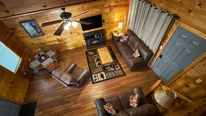 Interior acogedor de una cabaña con suelo de madera, ventilador de techo, chimenea, sofá, mesa de centro y televisor.