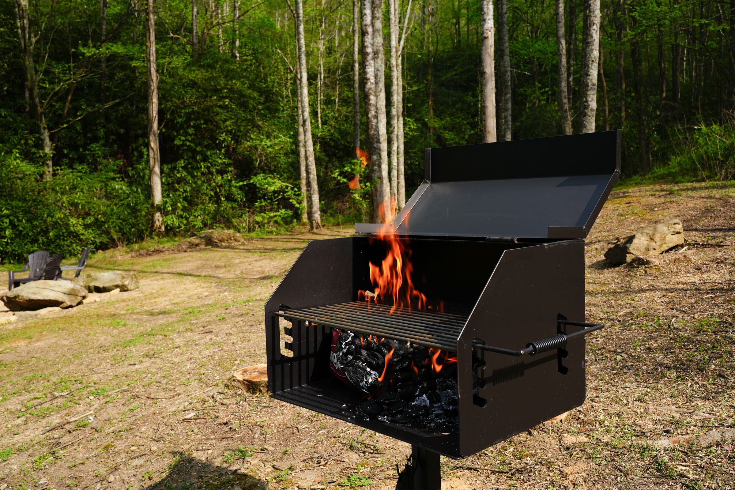 A black barbecue grill with flames, set in a forested outdoor area.