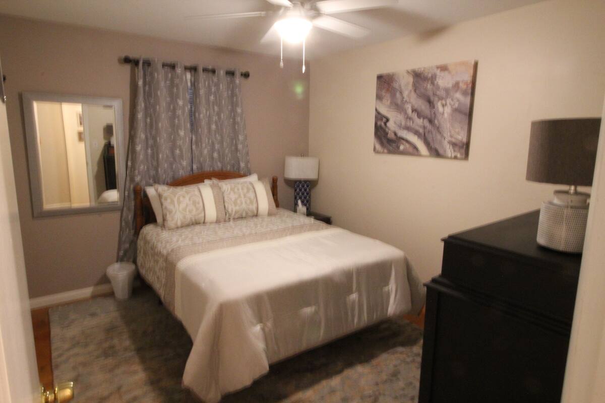 A bedroom with a bed, a ceiling fan, a nightstand with a lamp, a dresser, a trash can, and a painting on the wall.
