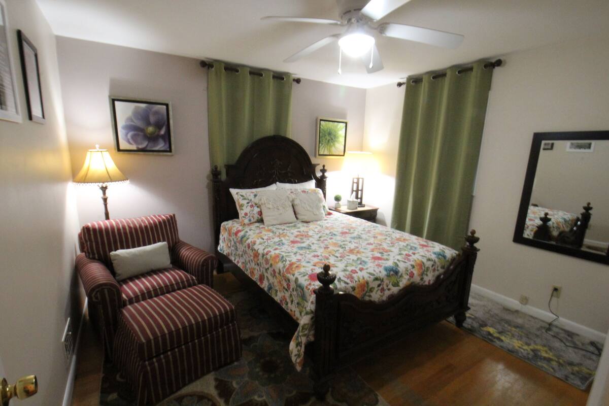 A bedroom with a bed, a chair, a nightstand, a lamp, a ceiling fan, and a mirror.