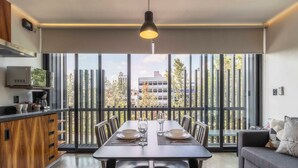 Una cocina moderna con microondas, cafetera y armarios. Hay un comedor con una mesa preparada para dos personas, sillas y un ventanal con vistas a edificios y árboles.