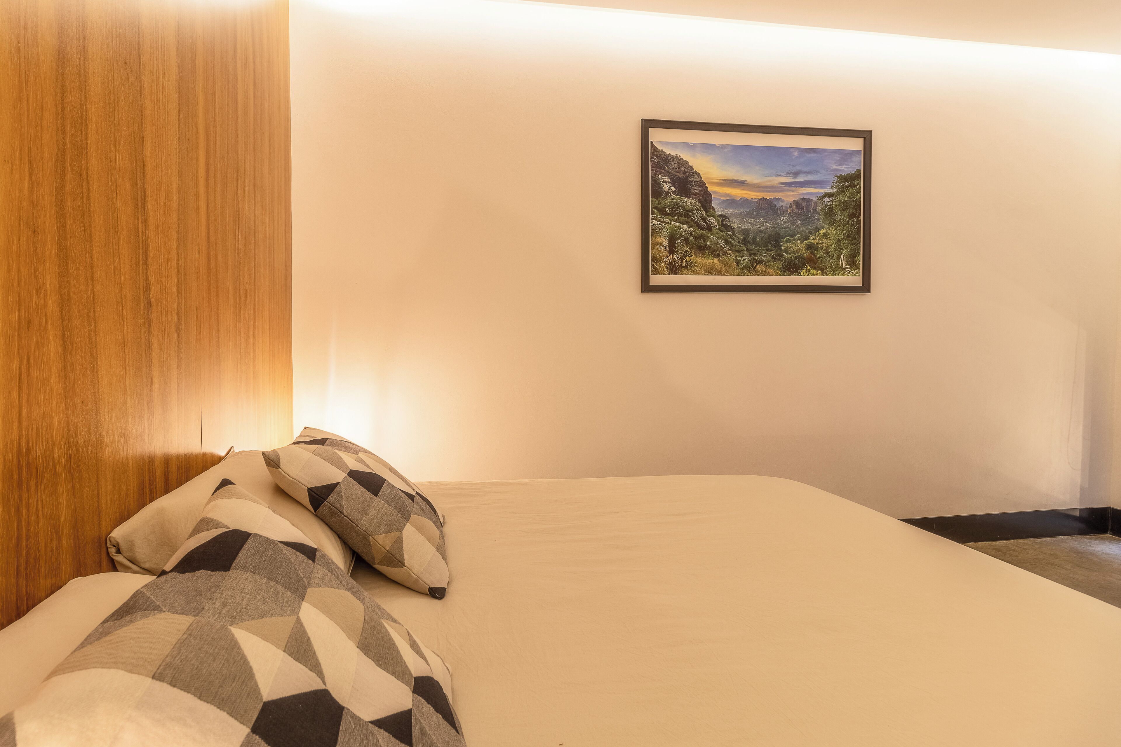 A neatly made bed with patterned pillows, a framed landscape painting on the wall, and a wooden headboard.