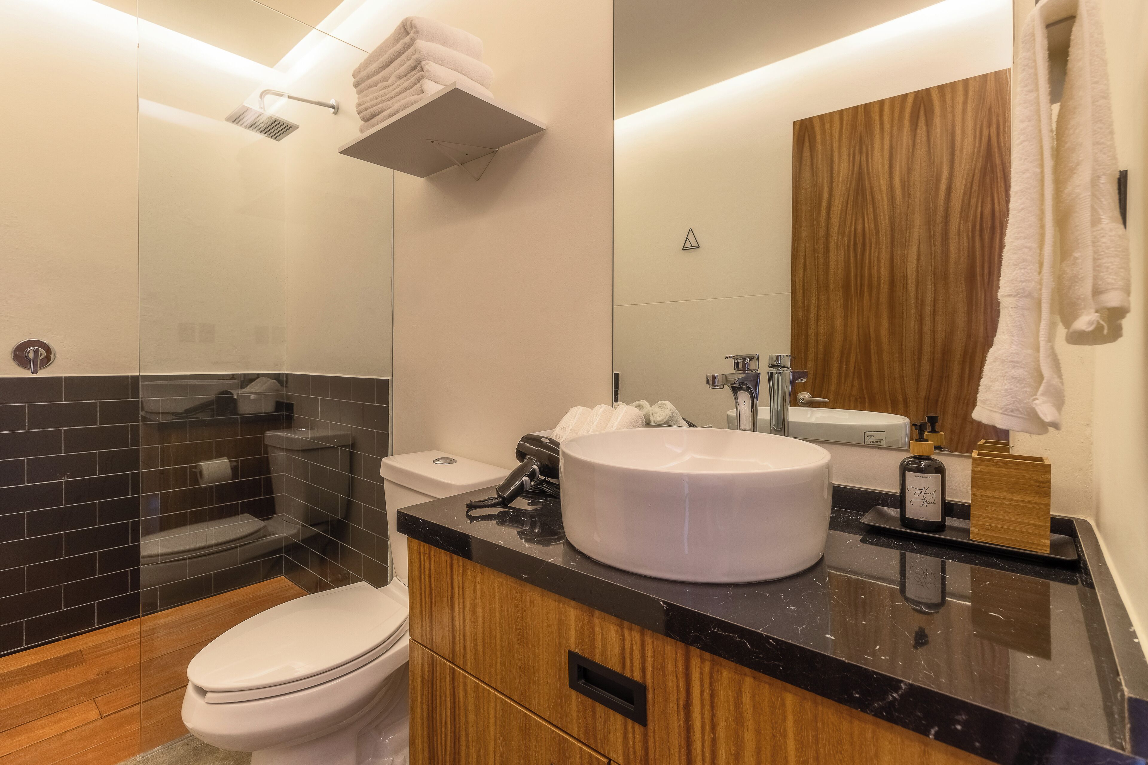 Une salle de bain moderne avec une douche aux carreaux noirs, des toilettes blanches, un meuble-lavabo en bois avec un lavabo blanc et un grand miroir.