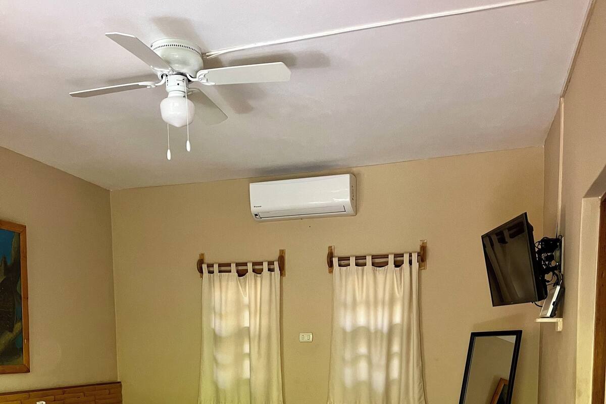 A hotel room with a bed, a ceiling fan, an air conditioning unit, a mirror, and a television mounted on the wall.