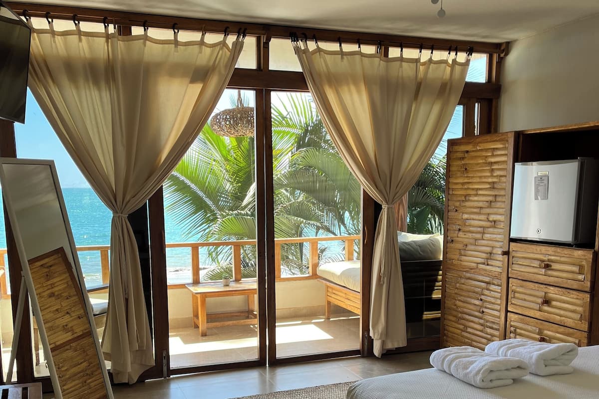 A bedroom with a bed, a wooden wardrobe, a ceiling fan, and a view of the ocean through large windows.