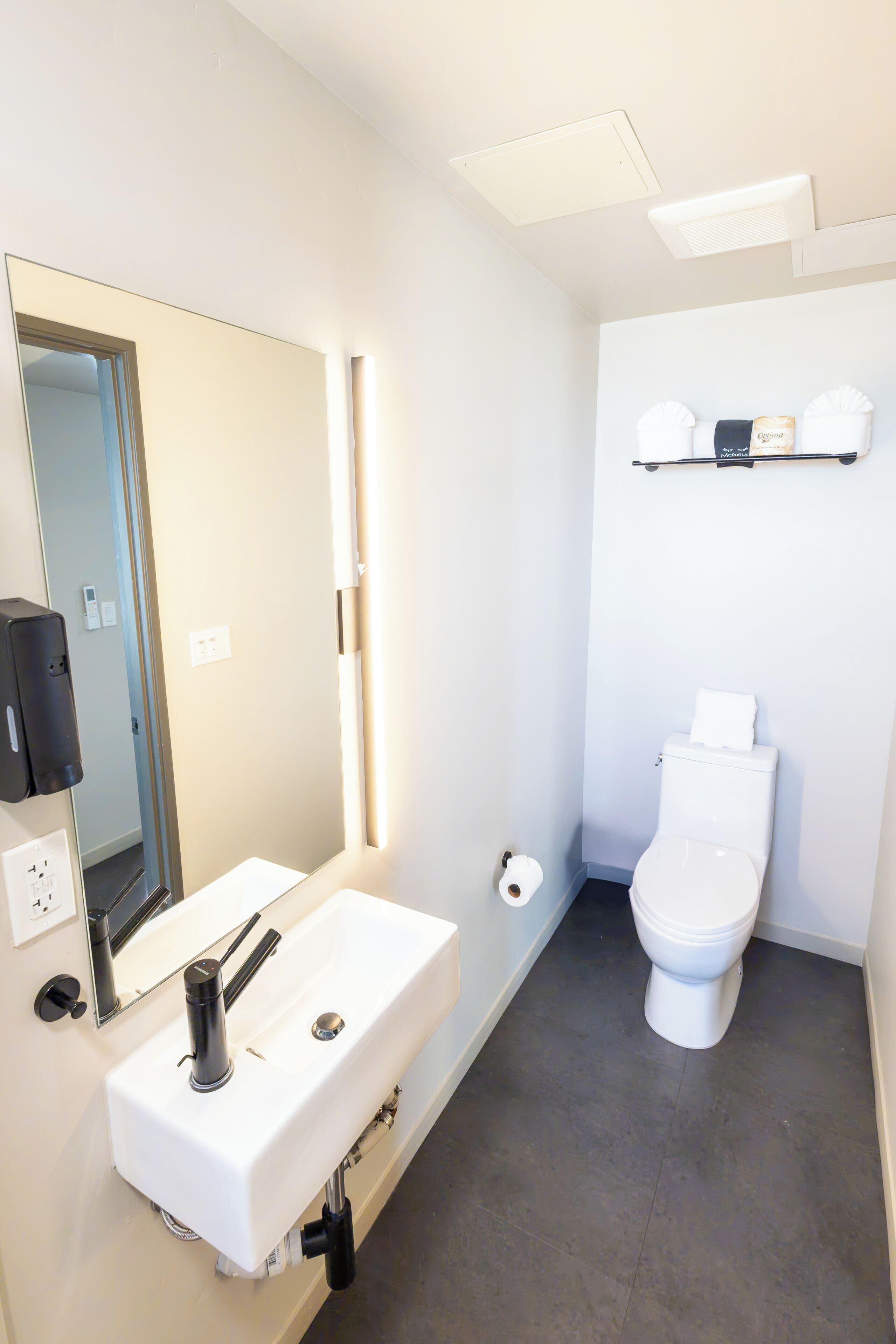 Un baño moderno con lavamanos blanco, espejo y retrete. Hay un perchero sobre el retrete y un dispensador de jabón en la pared.