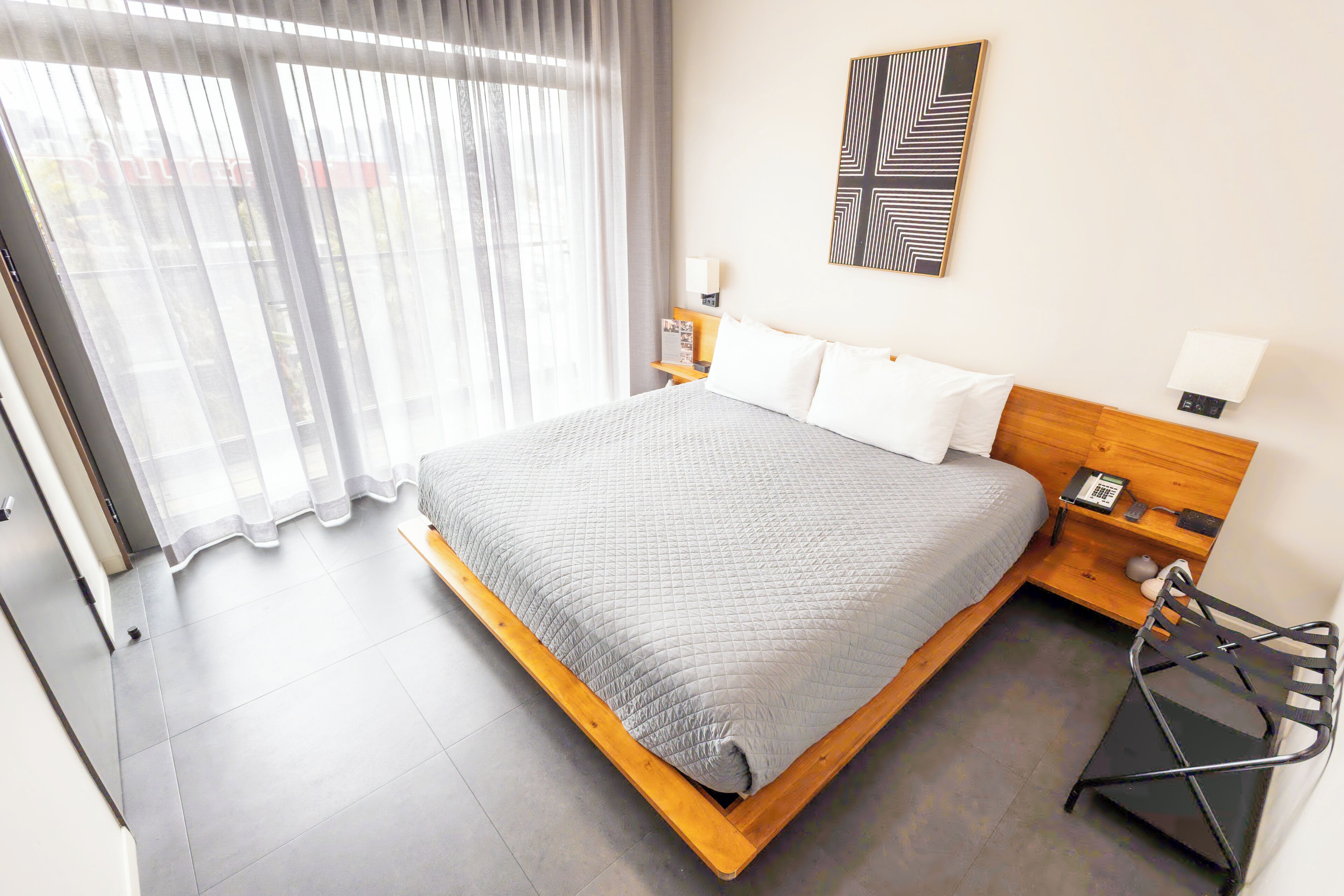 Une chambre d’hôtel avec un grand lit, une tête de lit en bois, deux tables de chevet avec des lampes, un téléphone fixé au mur et une fenêtre avec des rideaux légers.