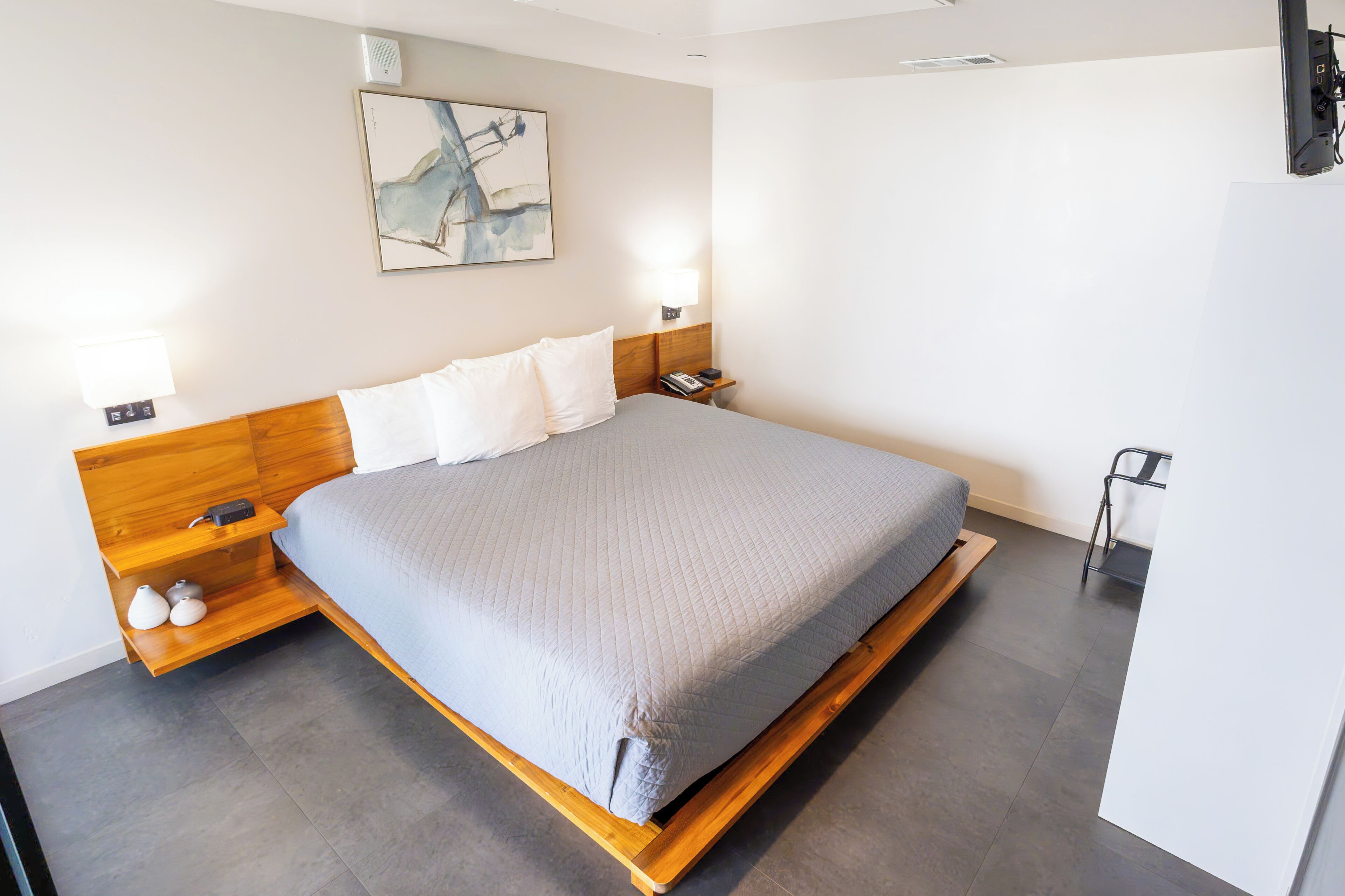 A modern hotel room with a large bed, bedside tables, a nightstand, a wall-mounted light, and a painting.