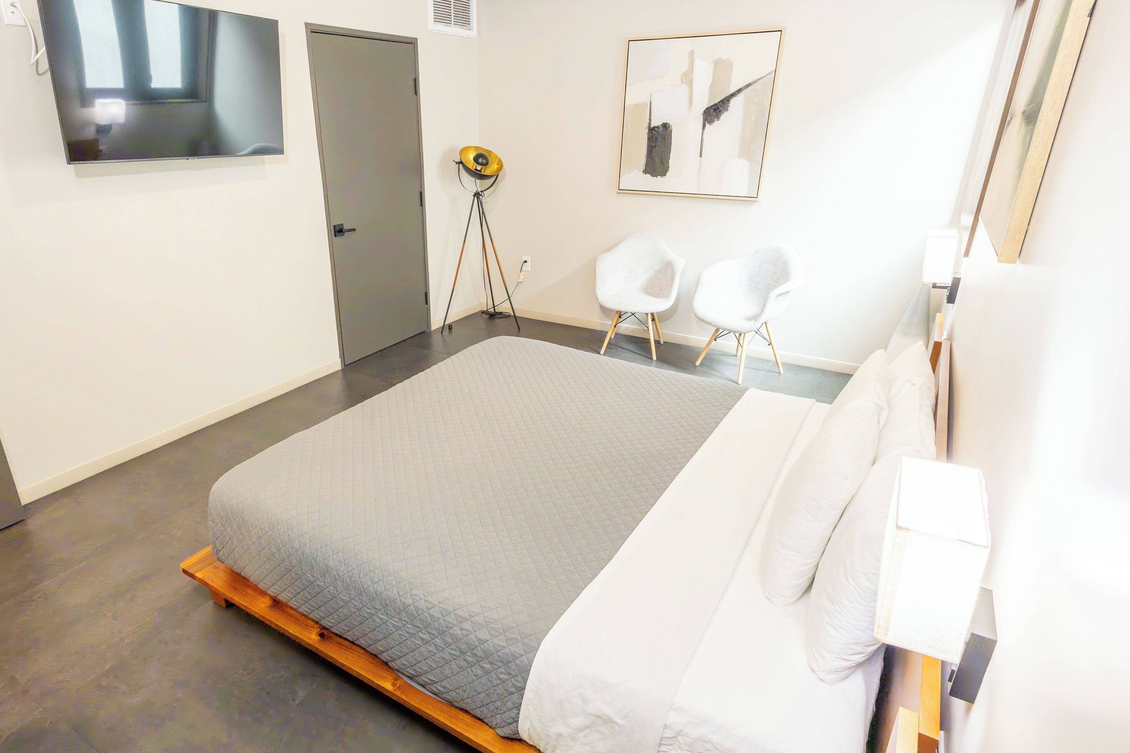 Une chambre à coucher moderne avec un grand lit, deux chaises blanches, un lampadaire sur trépied et un tableau accroché au mur.