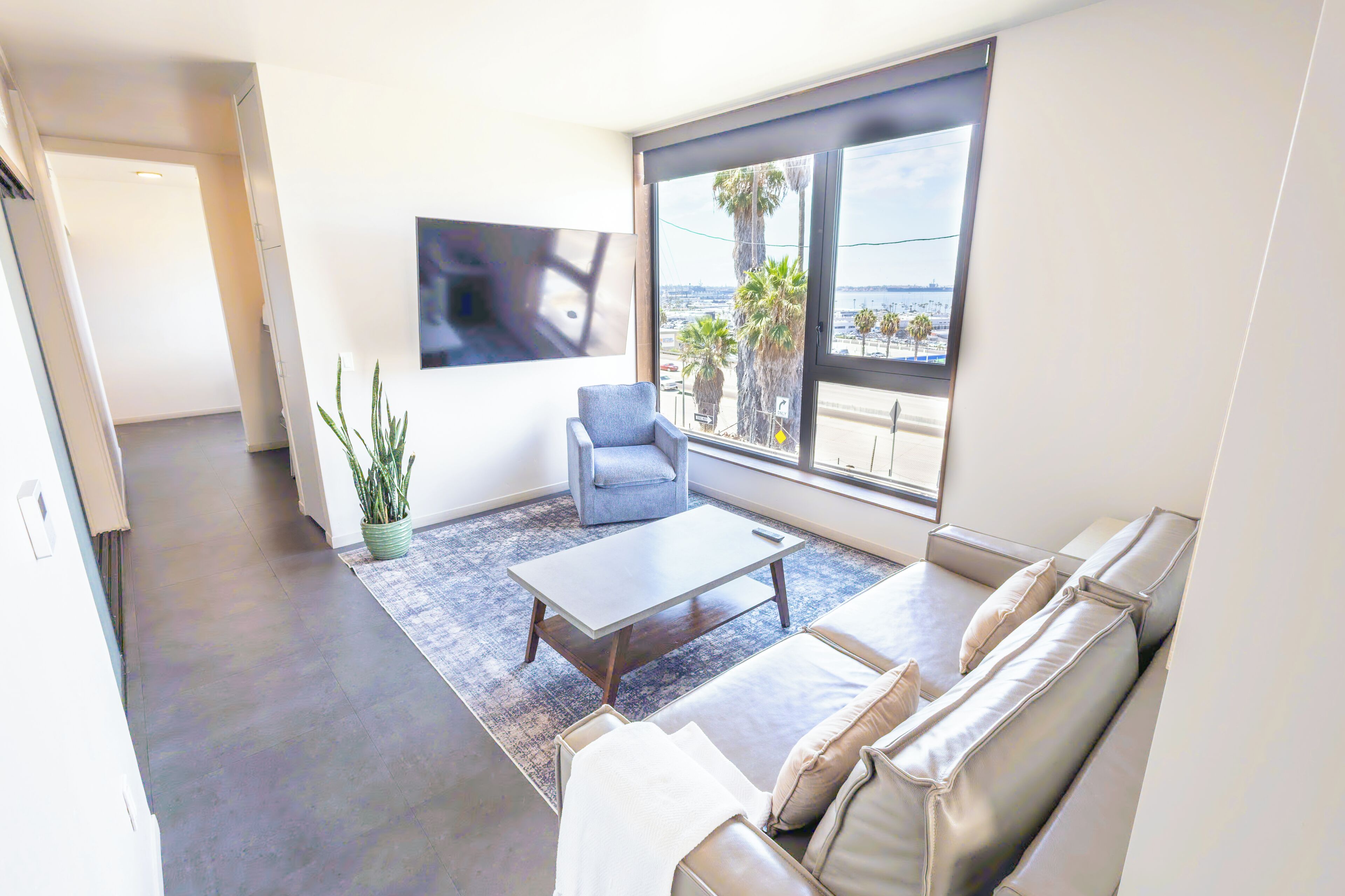 A modern living room with a sofa, a coffee table, a chair, a large window with a view of palm trees and a body of water, and a mounted TV.