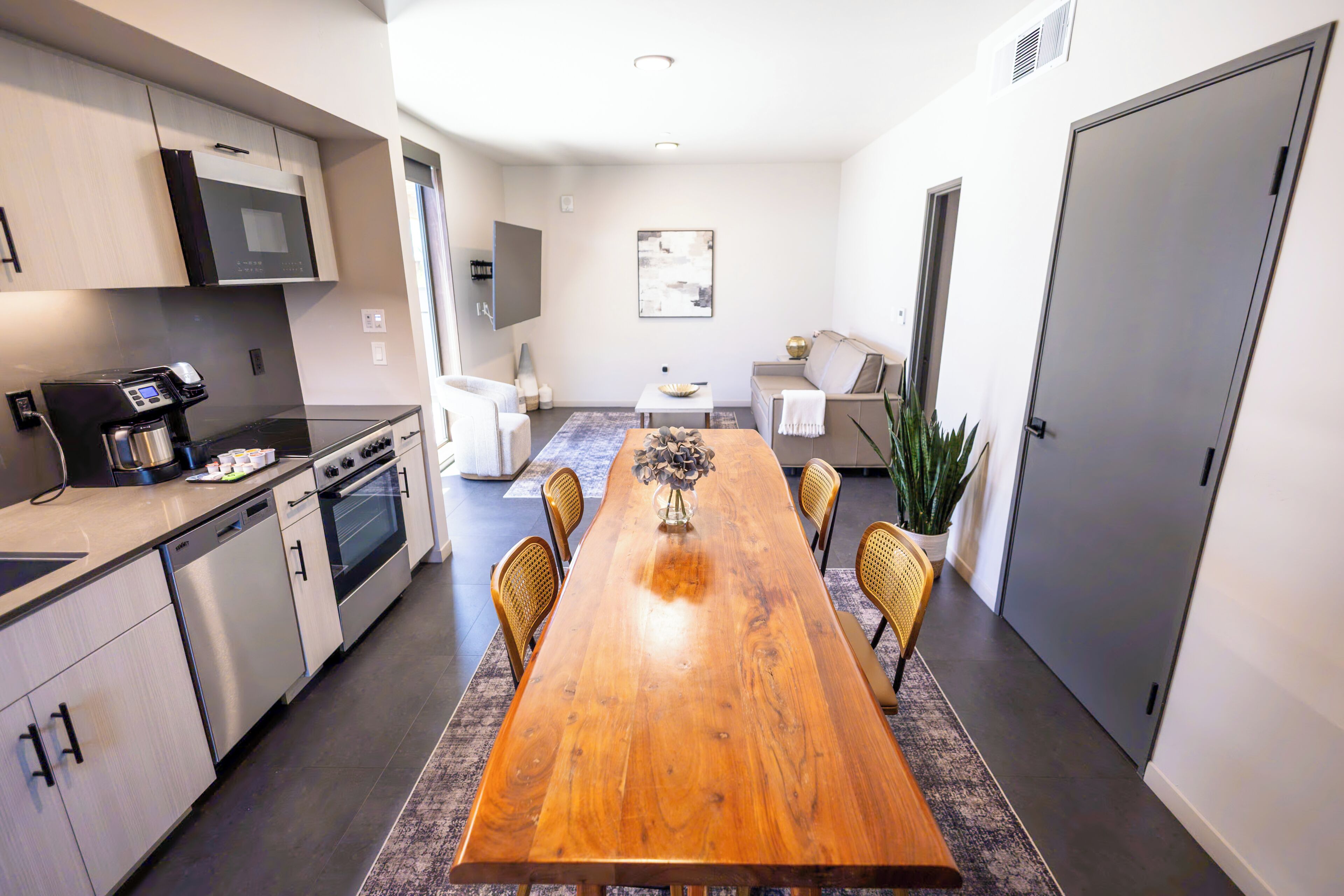 A modern kitchen with built-in appliances, a dining area with a wooden table and chairs, and a living space with a sofa and a plant.
