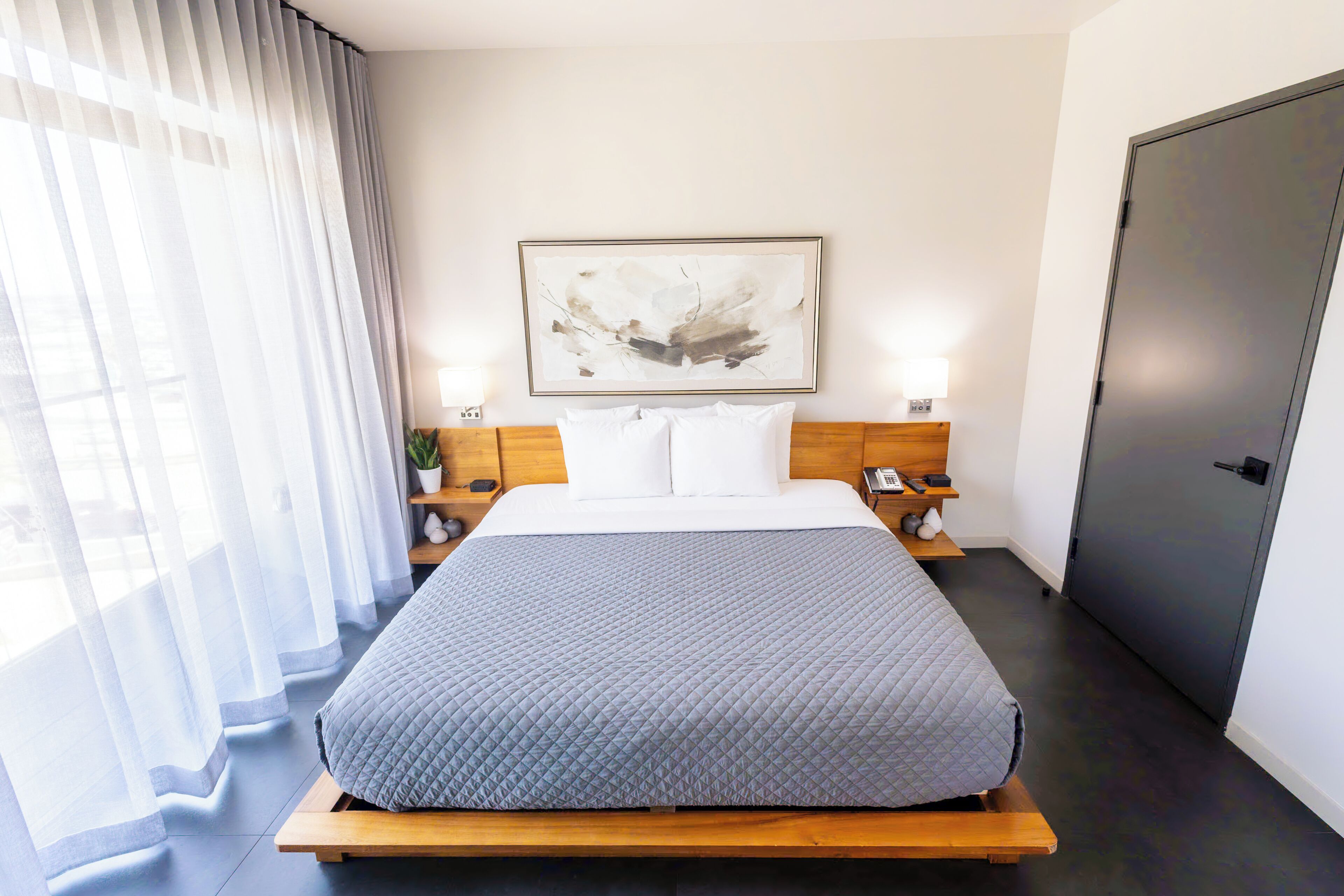 A hotel room with a bed, bedside tables, a painting, a door, and a window with curtains.