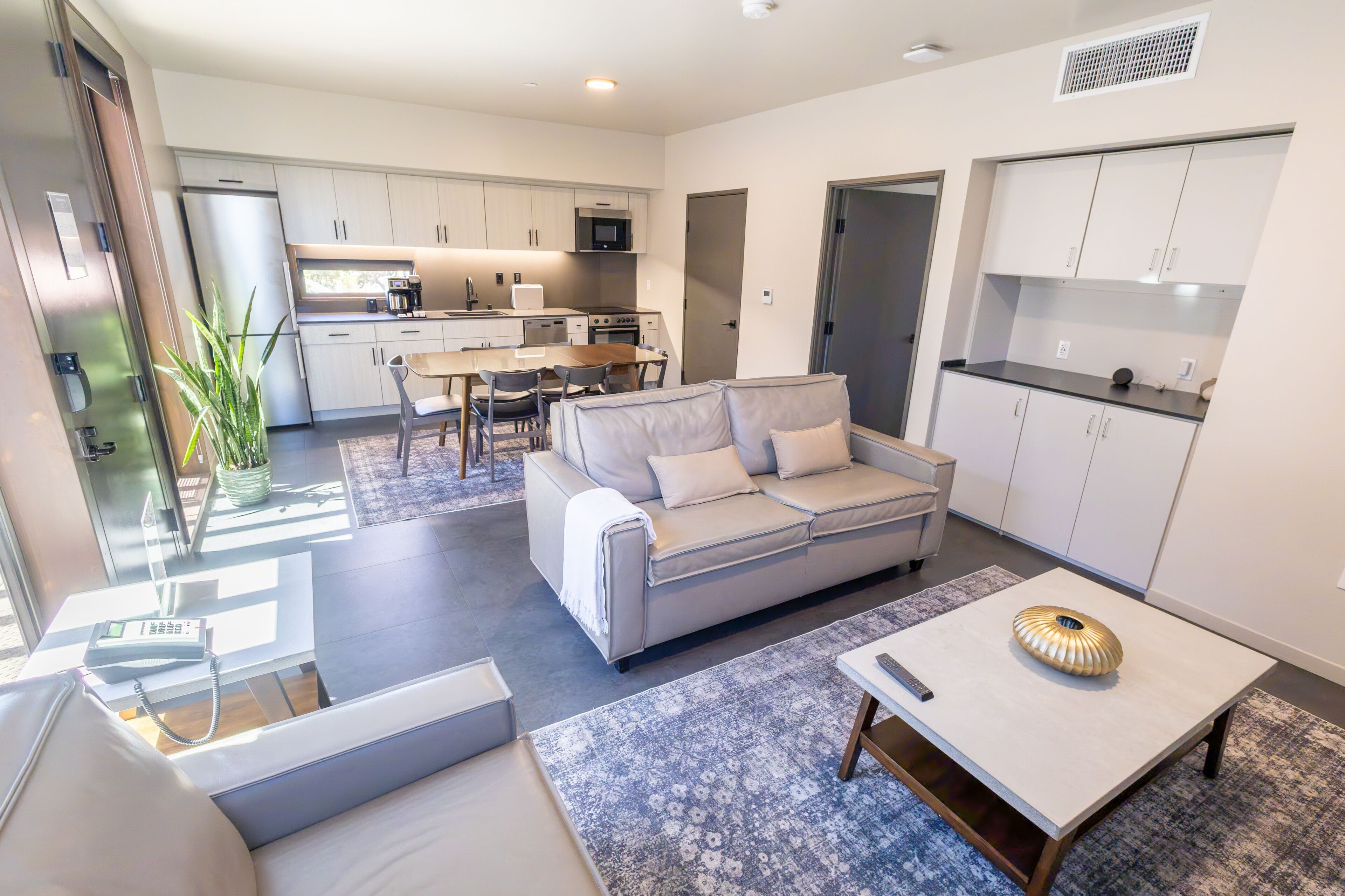 A modern kitchen with stainless steel appliances, a dining area with a table and chairs, a sofa with cushions, and a small coffee table.
