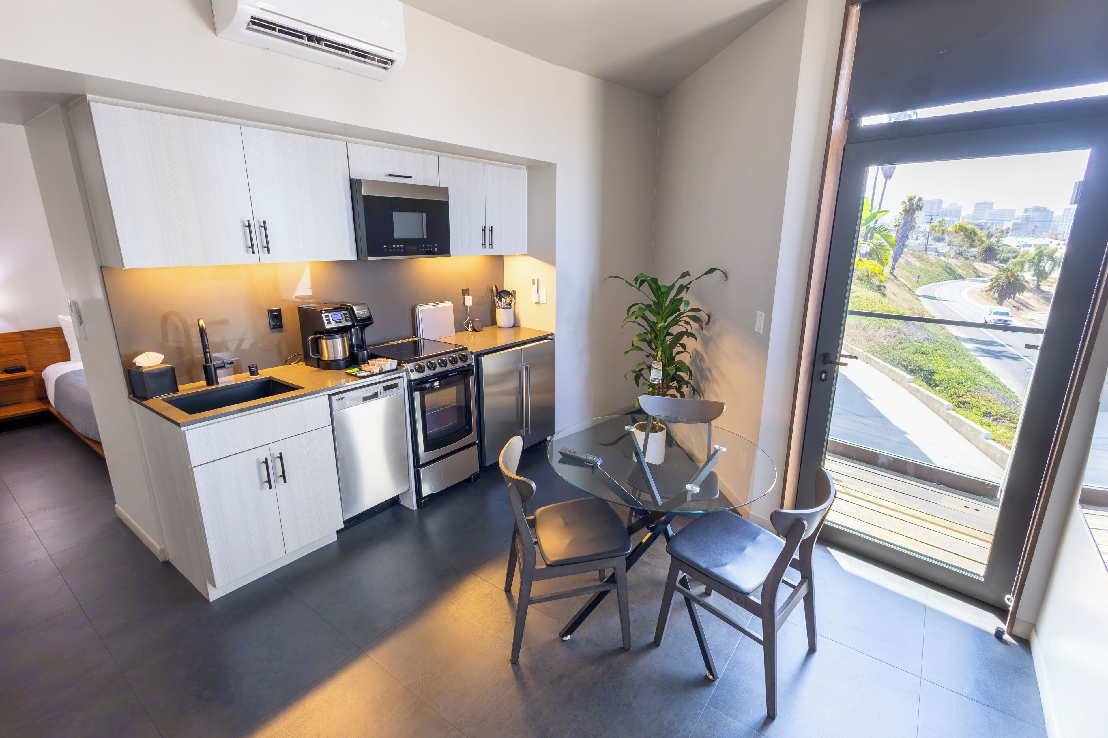 A modern kitchen with a built-in microwave, oven, and dishwasher. There's a dining area with a glass table and chairs, and a view of a cityscape through a large window.