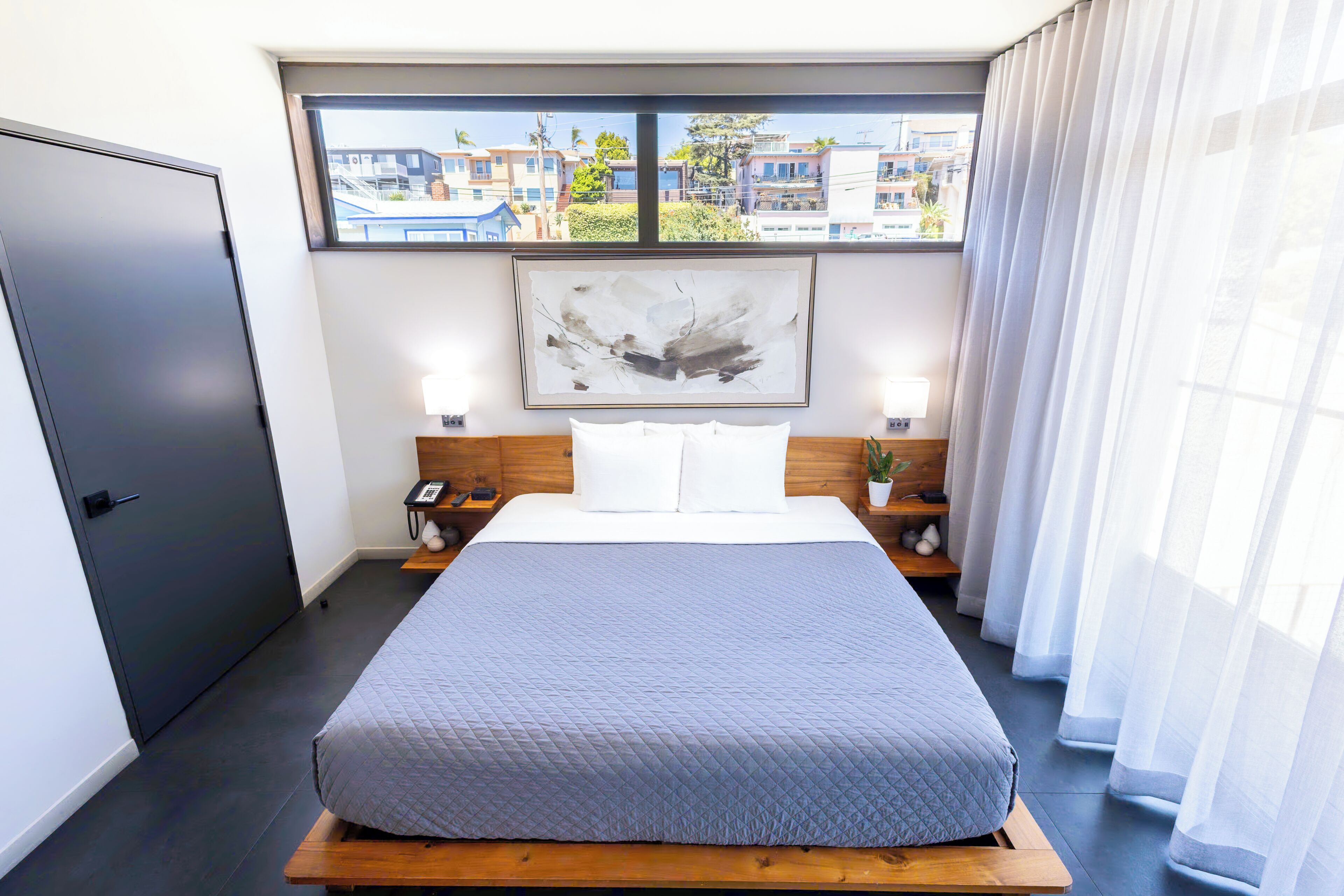 A modern hotel room with a large bed, a wooden headboard, a painting above the bed, a bedside table with a lamp, and a view of buildings through the window.