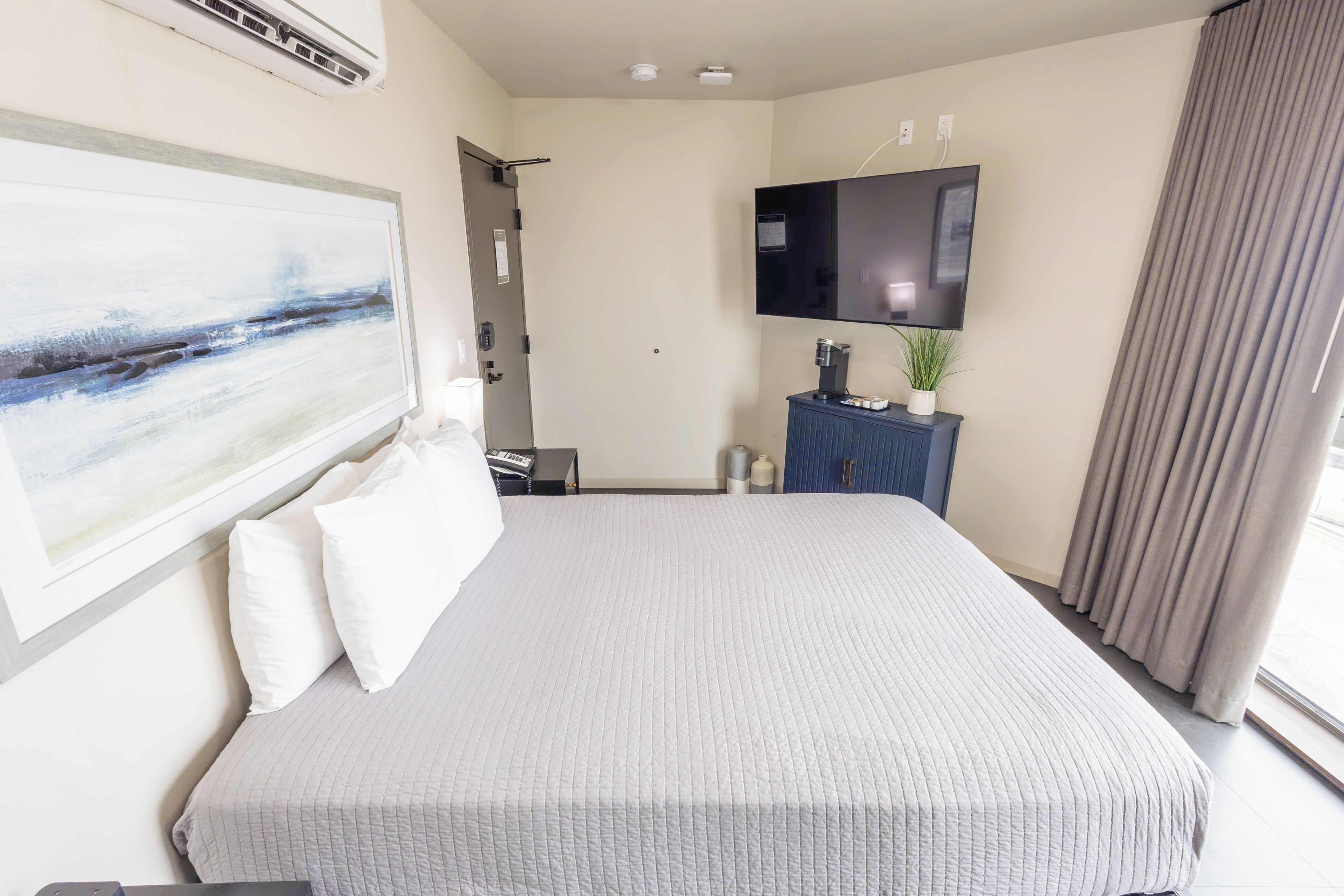 A hotel room with a bed, a painting, a television, a minibar, and a window with curtains.