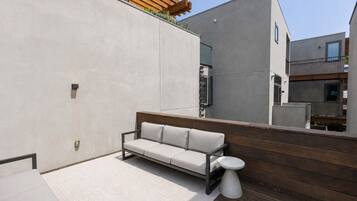 A modern outdoor seating area with a sofa and a small table, a wooden deck, and a building with a minimalist design.
