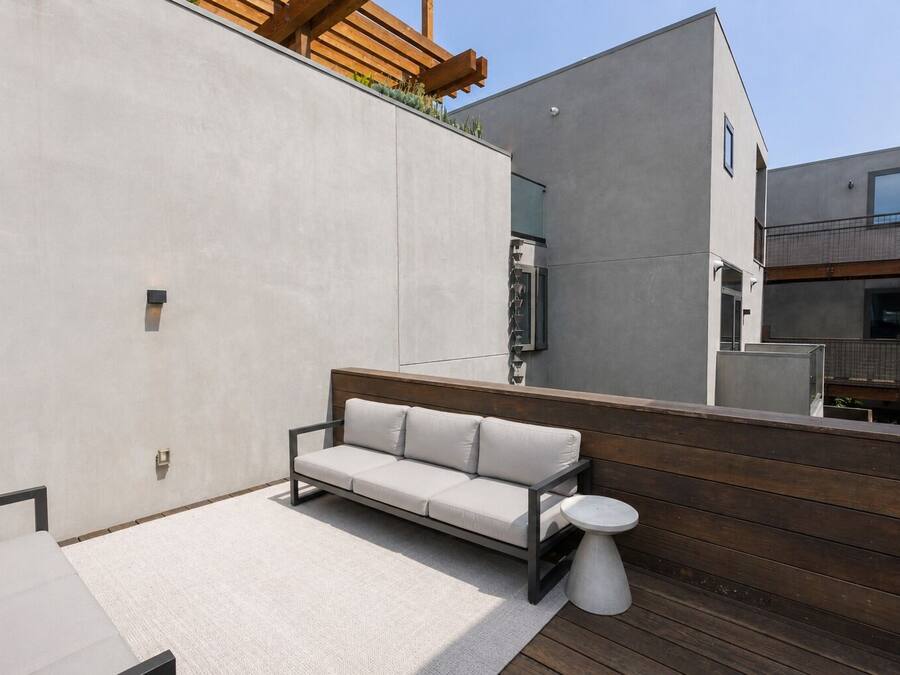 A modern outdoor seating area with a sofa and a small table, a wooden deck, and a building with a minimalist design.