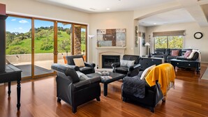 A spacious living room with a fireplace, leather sofas, and a view of a green landscape through large sliding glass doors.