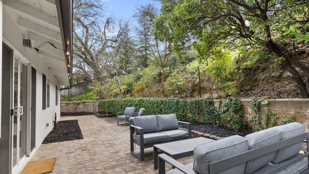 Un patio exterior con suelo de ladrillo, mobiliario exterior gris y un seto bien cuidado.