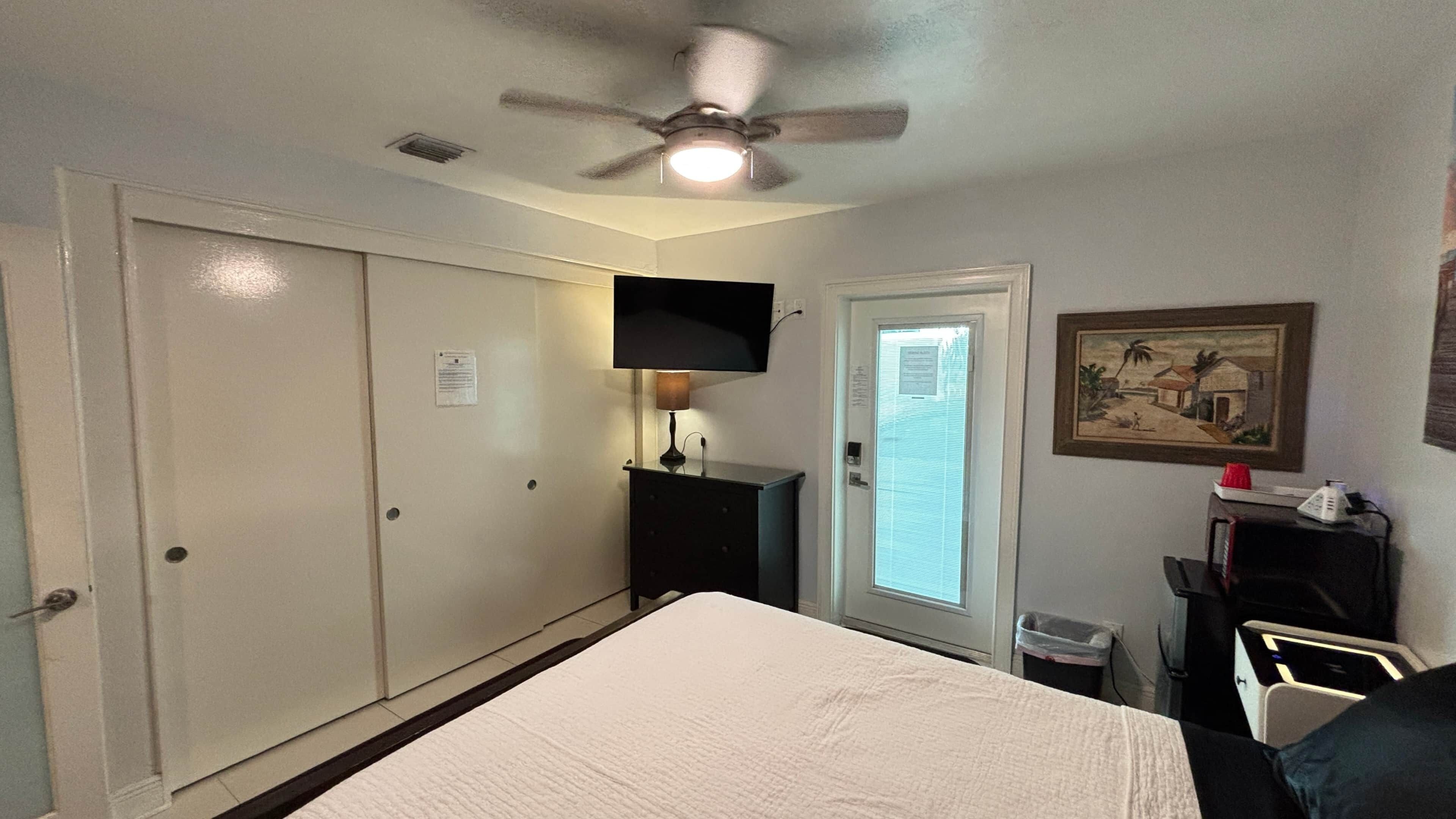 A hotel room with a bed, a ceiling fan, a TV mounted on the wall, and a door with a window.