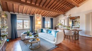 A modern living room with a wooden ceiling, a white sofa with blue cushions, a dining area with a table set for four, and a view of the city through large windows. - De Nobili Penthouse in Lucca (Lucca)