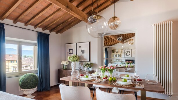 A dining area with a wooden table set for four, white chairs, and a view of the city through large windows. - De Nobili Penthouse in Lucca (Lucca)