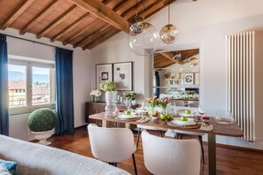A dining area with a wooden table set for four, white chairs, and a view of the city through large windows. - De Nobili Penthouse in Lucca (Lucca)