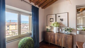 A room with a large window, wooden ceiling, a round green plant, a wooden cabinet with vases, and framed artwork. - De Nobili Penthouse in Lucca (Lucca)