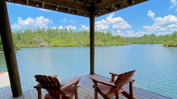 Una terrassa de fusta amb dues cadires Adirondack i una taula, amb vista a un llac tranquil i una riba boscosa.