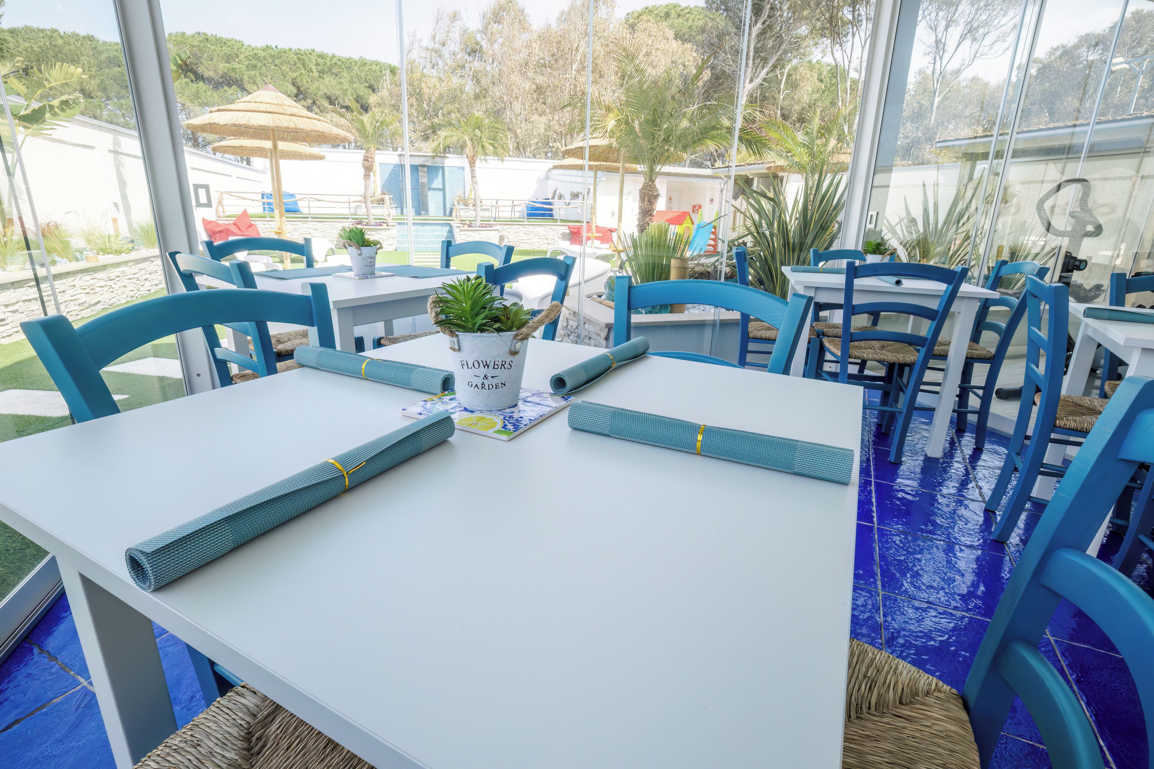 Comedor con mesas blancas y sillas azules, una planta en maceta y vistas a la piscina y la vegetación exterior.