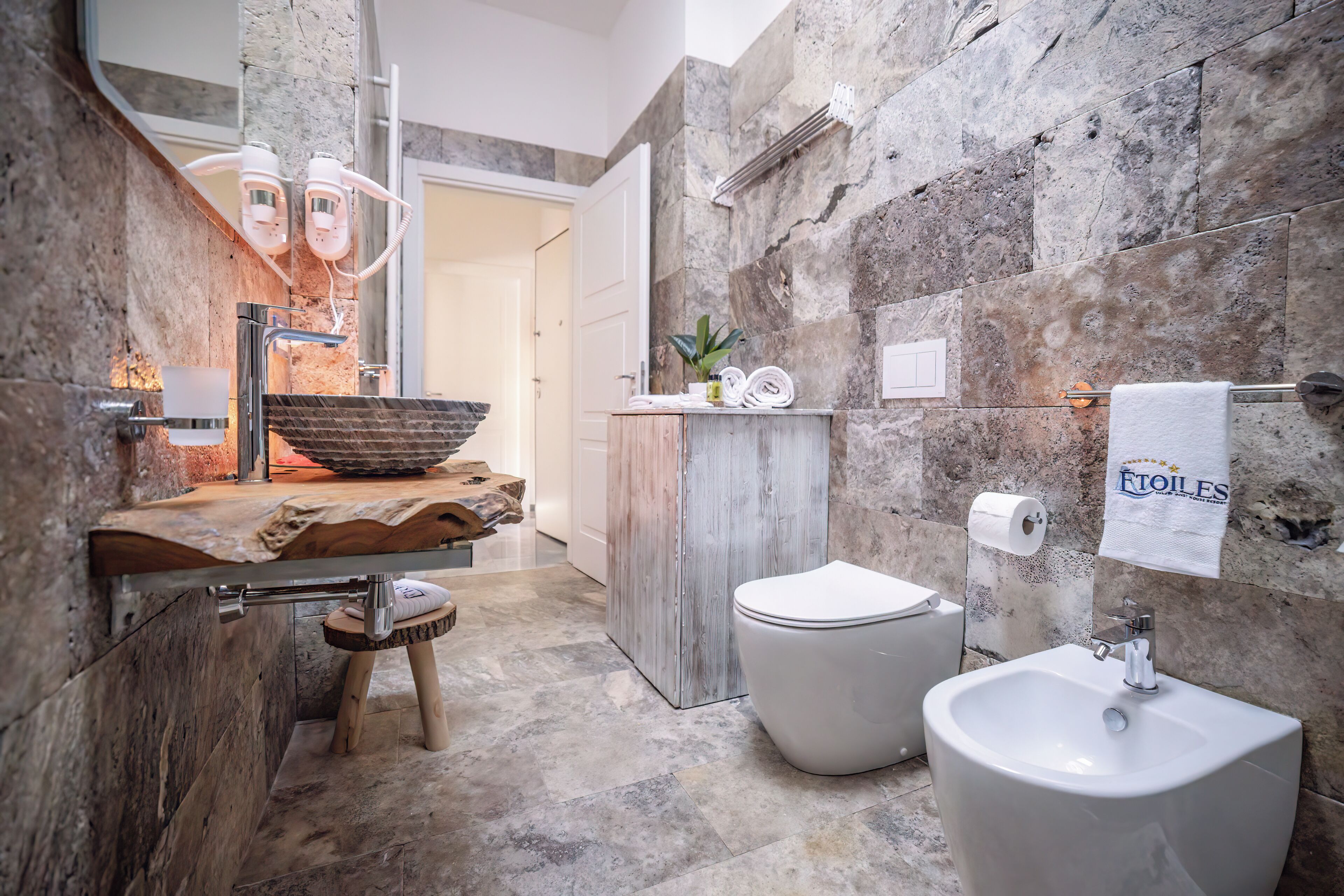 A modern bathroom with a stone wall, a wooden vanity, a white sink, and a bidet.