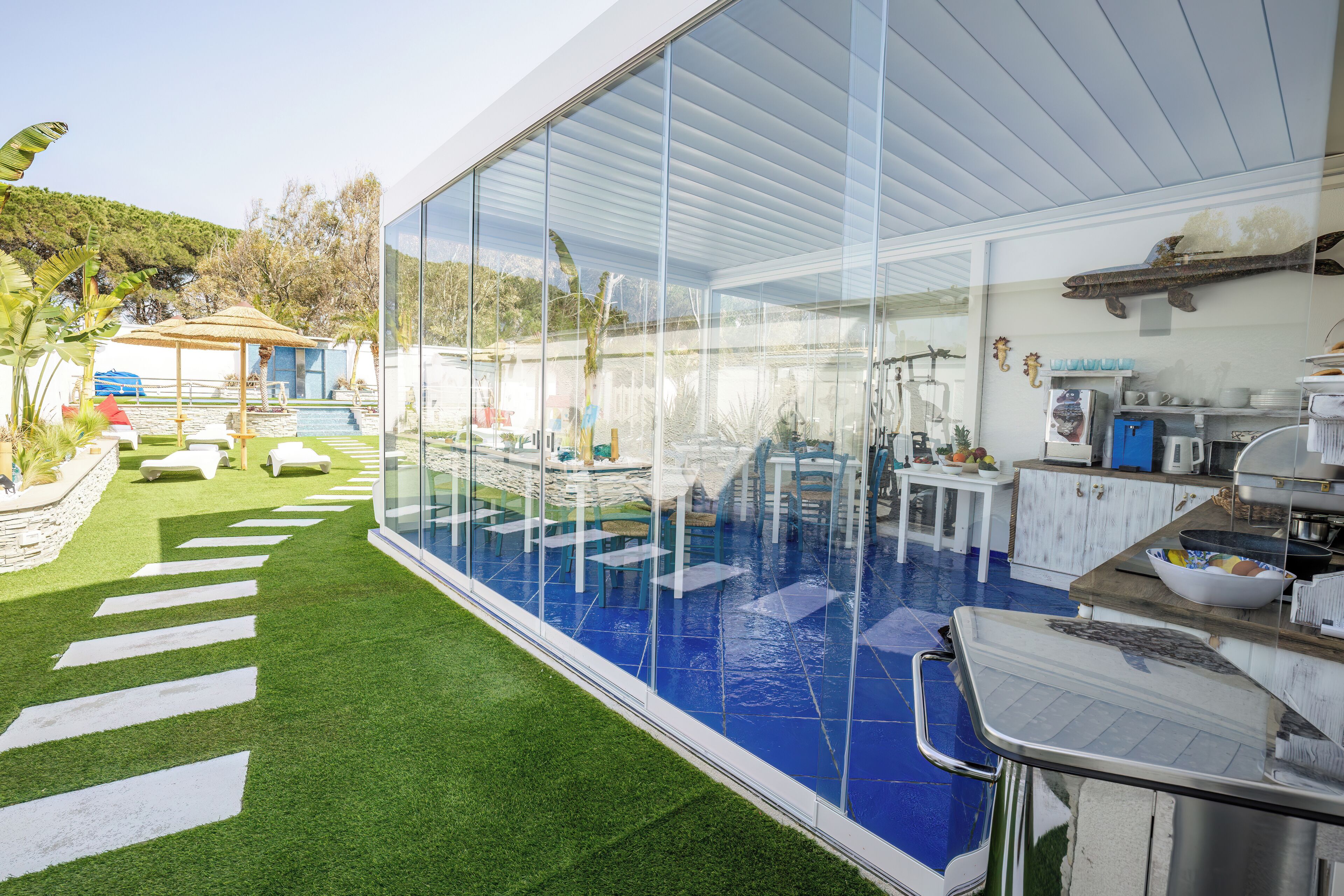 Un espace piscine extérieur moderne doté de murs en verre, d’un coin repas et d’une cuisine visible à travers le verre.