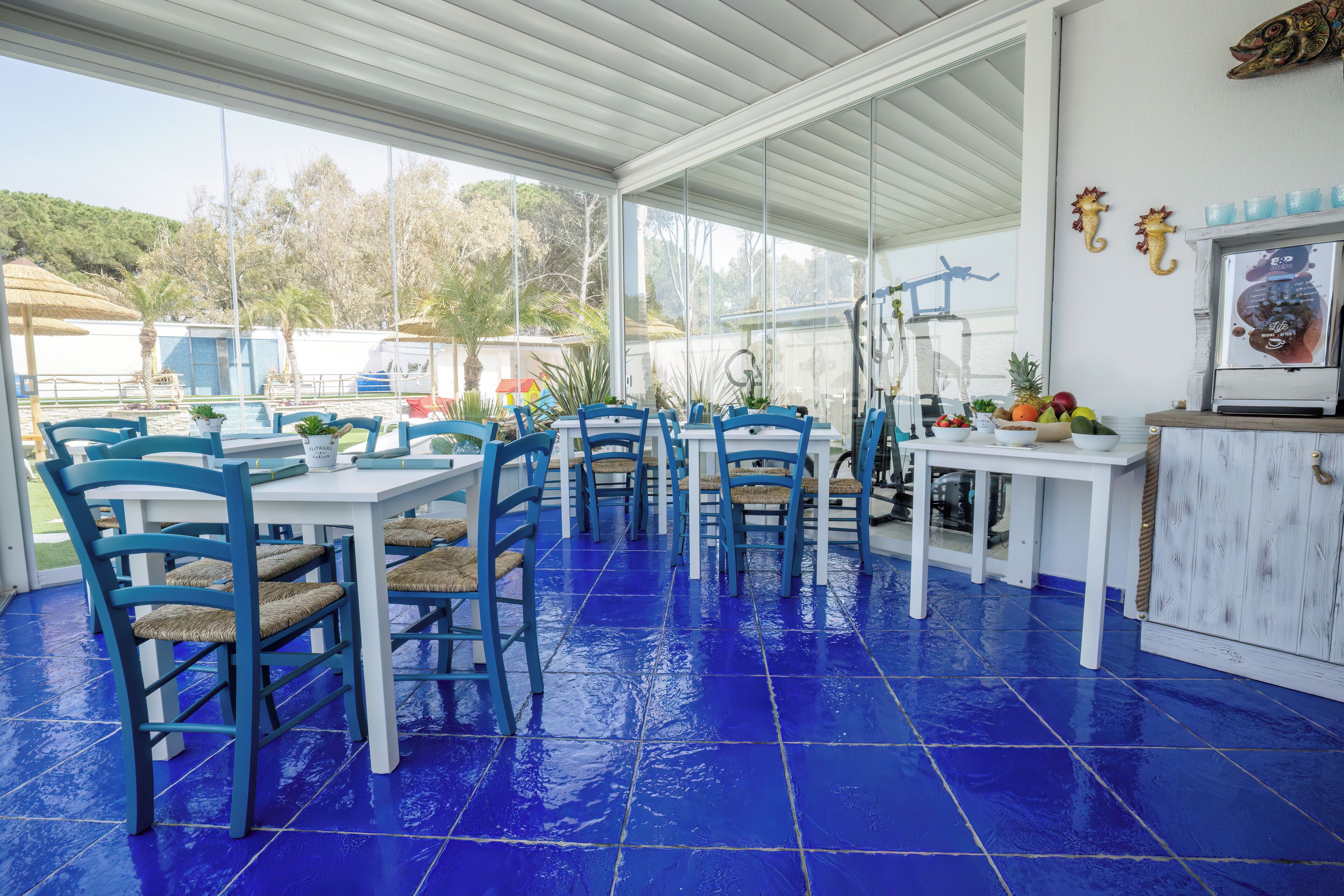 Un espacio comedor al aire libre techado con sillas y mesas azules, un mueble blanco con adornos, y vistas del entorno.