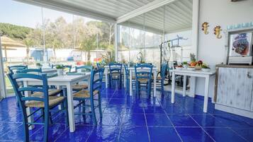 Un espacio comedor al aire libre techado con sillas y mesas azules, un mueble blanco con adornos, y vistas del entorno.
