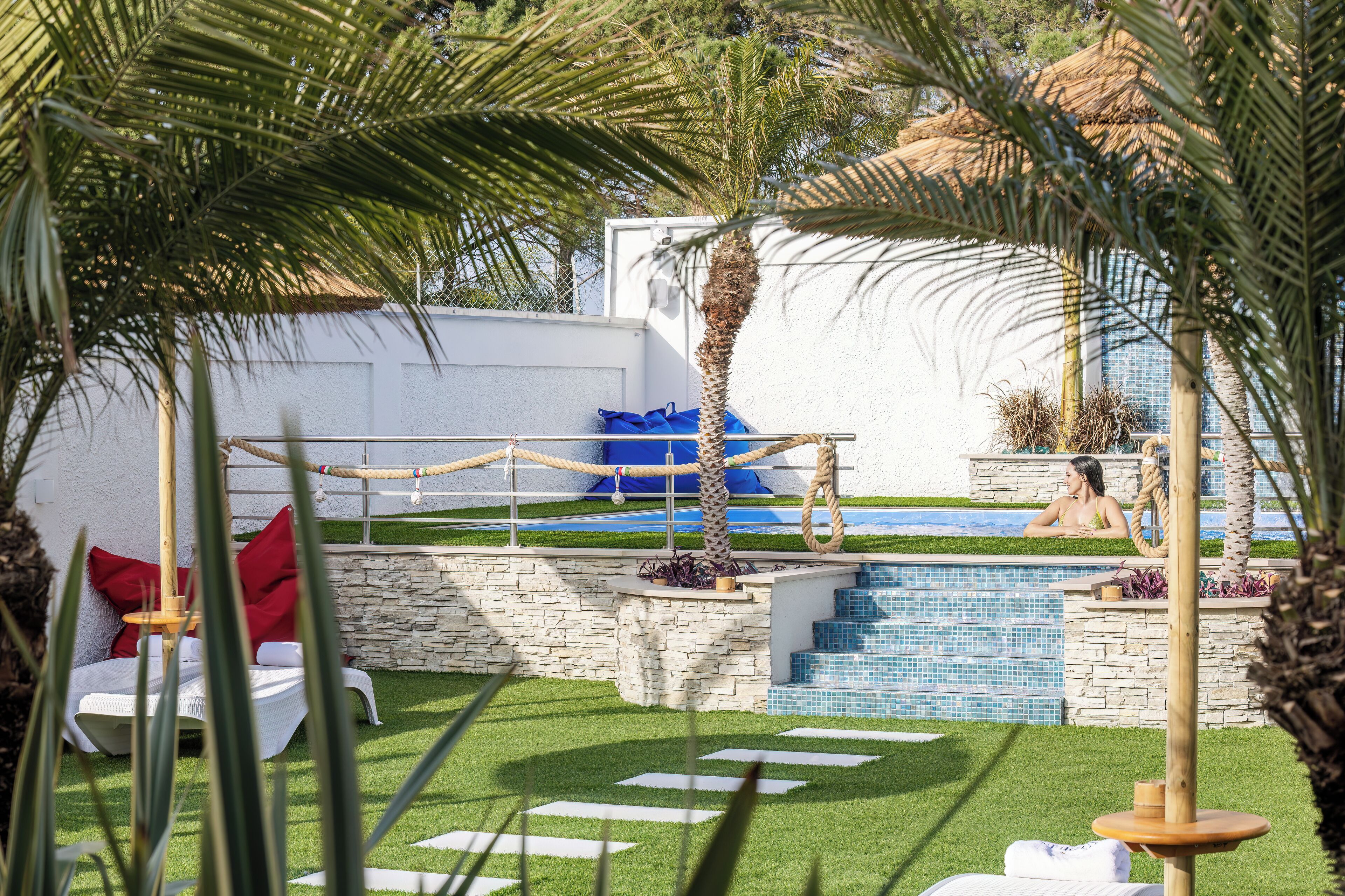 Un espace piscine agrémenté d’un mur en pierre, d’une petite piscine et d’un coin salon avec une chaise blanche et des coussins rouges.