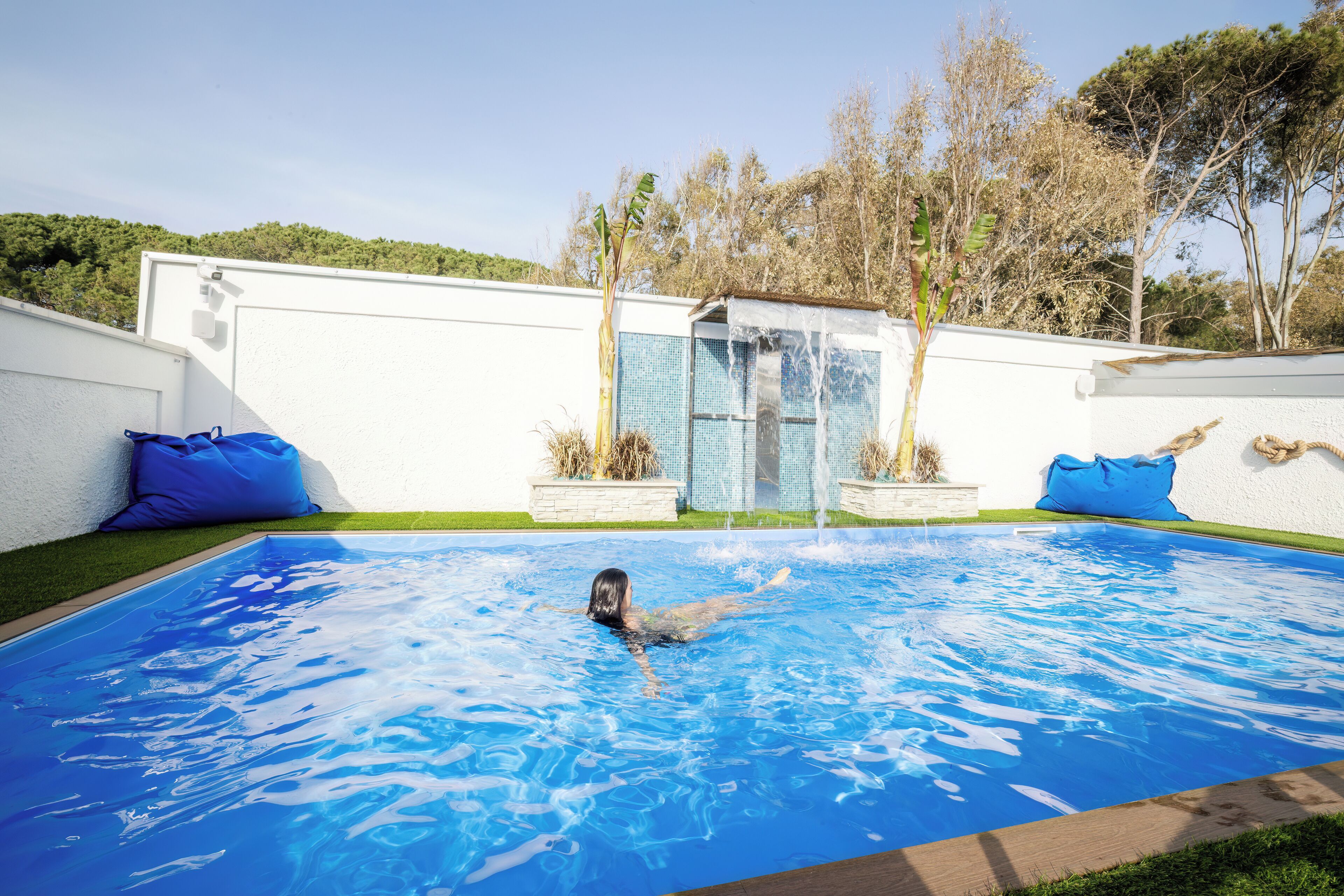 Piscina com uma pessoa nadando, rodeada por um muro branco e almofadas azuis.