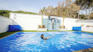 Piscina com uma pessoa nadando, rodeada por um muro branco e almofadas azuis.