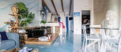 A spacious living area with a blue-tiled floor, a sofa, a glass coffee table, and a dining table with white chairs.
