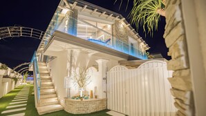 Una casa moderna de dos pisos con un balcón de vidrio, una escalera de piedra y un portón blanco.