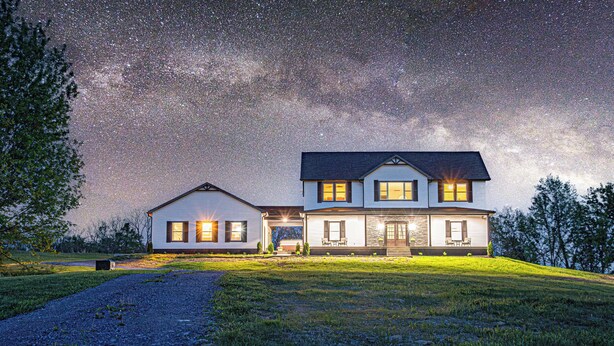 Uma casa de dois andares com varanda coberta e um jardim frontal bem iluminado sob um céu estrelado.