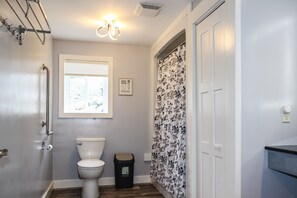 A bathroom with a toilet, a trash bin, a shower curtain, and a window with blinds.