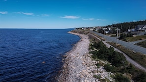 A coastal road with houses on the right, a rocky shoreline, and a clear blue sky.