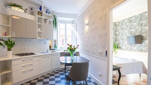 A modern kitchen with white cabinets, a black and white checkered floor, a dining table with a vase of flowers, and a view of a balcony with a TV. - Stilysh apartment in Rome close to cafes, restaurants, bus and metro. (Roma)