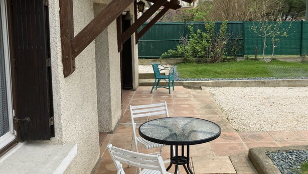 En uteplats med ett runt bord och två vita stolar, en träpergola och ett grönt staket i bakgrunden.