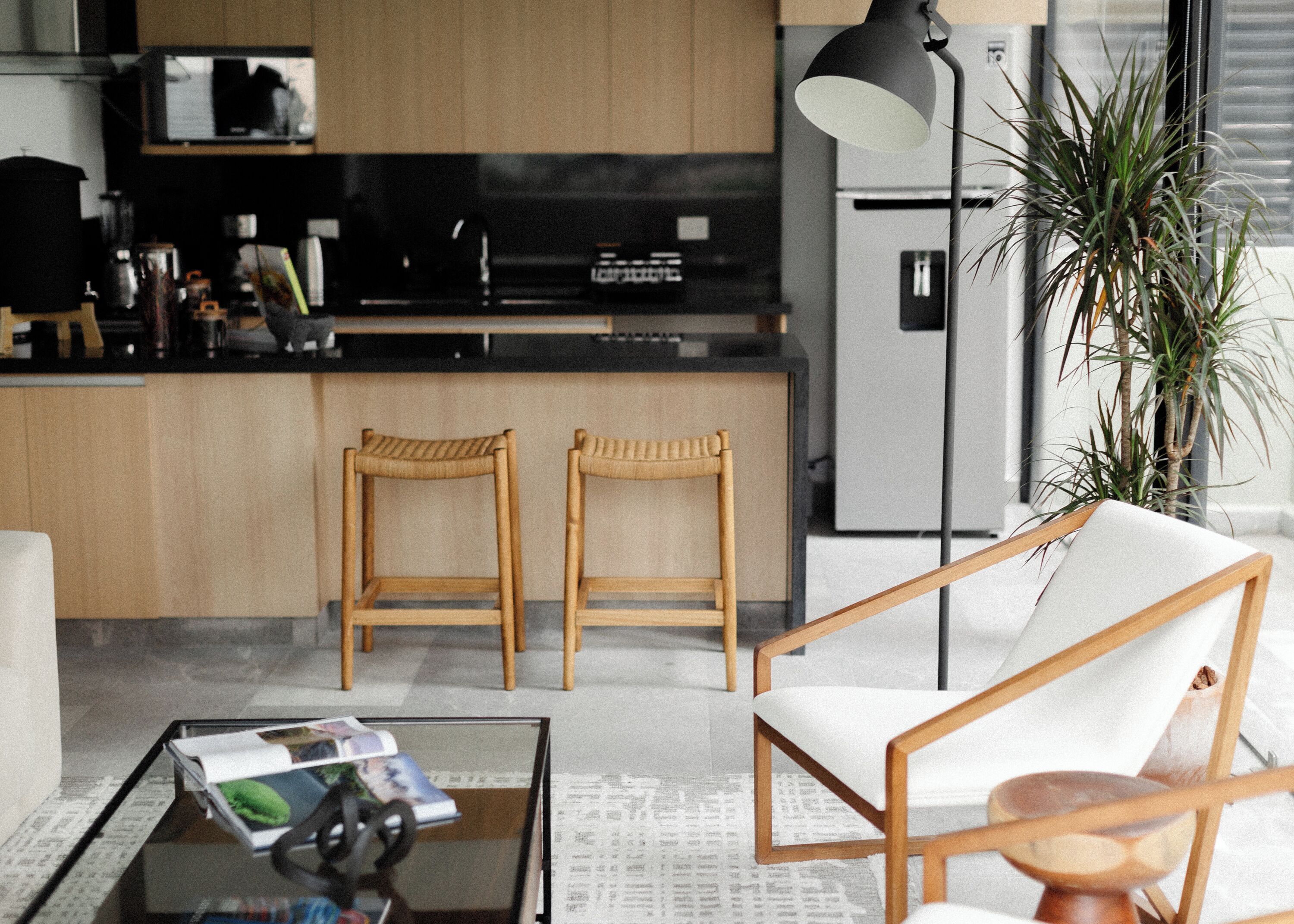 A modern kitchen with wooden stools, a dining table with magazines, and a large plant.