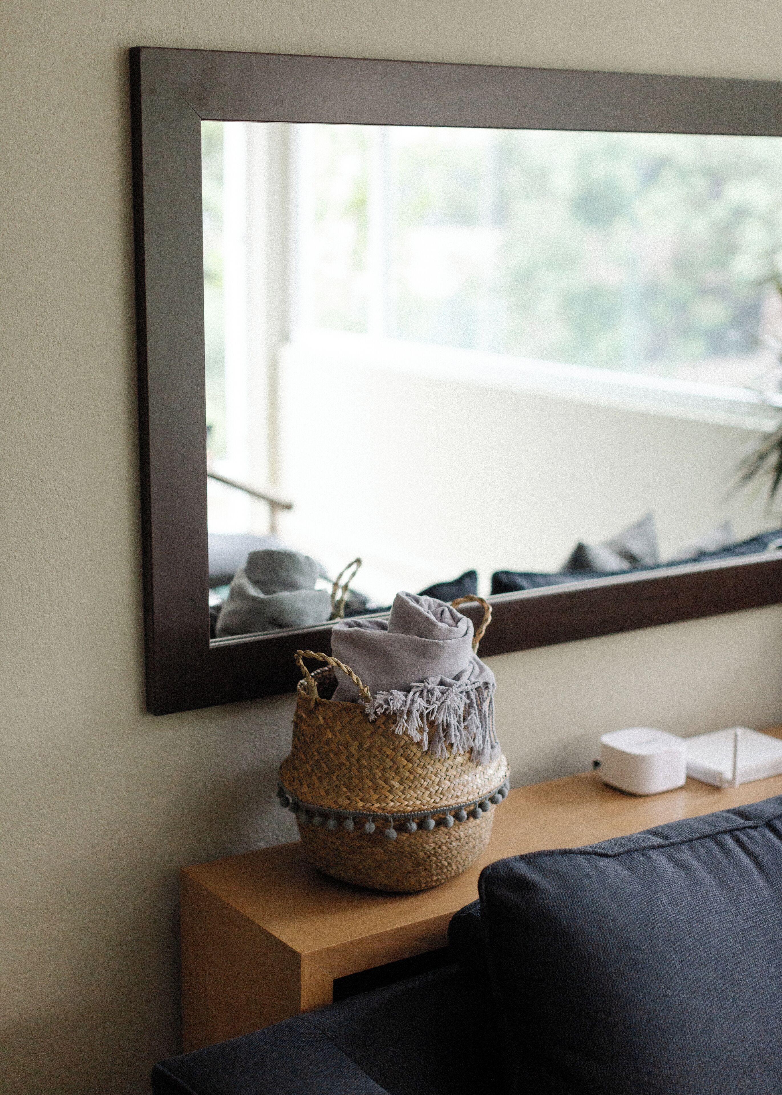 A room with a large mirror on the wall, a woven basket on a wooden side table, and a dark-colored sofa.