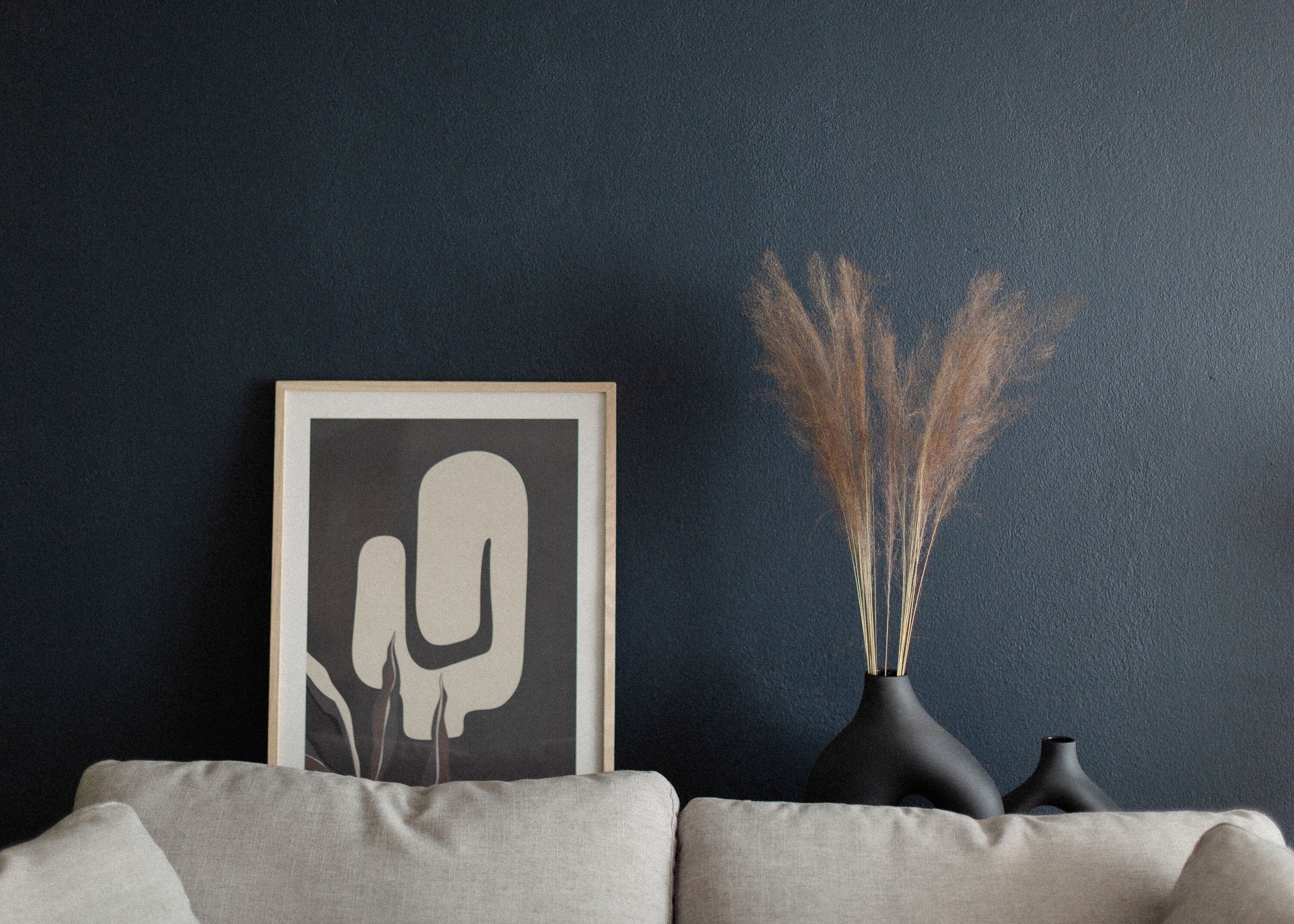 Et indrammet abstrakt kunstværk på en mørk væg, en vase med visne planter, og en sofa.